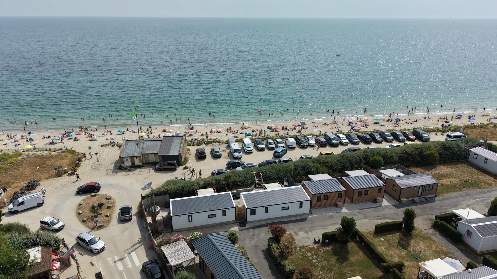 Vue aérienne : au premier plan des mobil-homes du camping, puis une haie, une allée où des voitures sont garées, la plage et l’océan