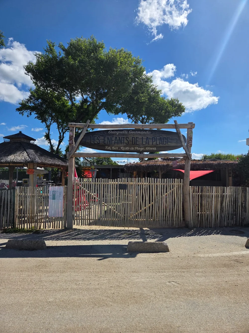 L’entrée de la guinguette Les Enfants de la Plage : l’enseigne en bois en forme de planche de surf au-dessus du portail en palissade de châtaignier, les paillotes au toit de chaume et le ciel bleu d’été