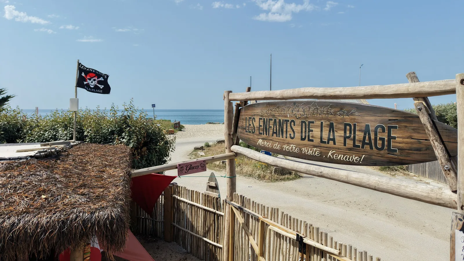 L’enseigne en bois de la guinguette, « Les Enfants de la Plage, merci de votre visite, Kenavo ! », au-dessus d’une palissade, avec le drapeau de pirate de la guinguette, le chemin de sable et la mer au fond