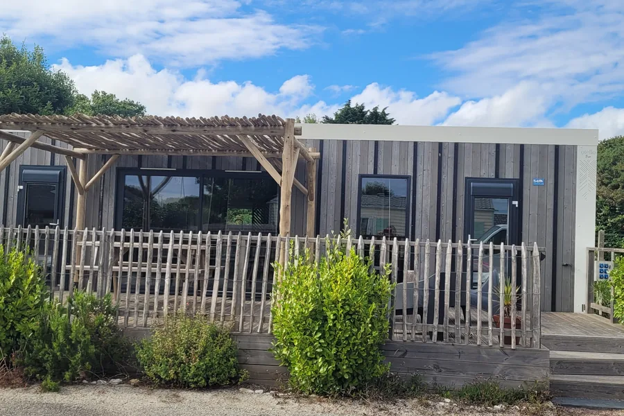 Le mobil-home Malibu, pergola en rondins et terrasse fermée par une ganivelle, devant des massifs d’arbustes