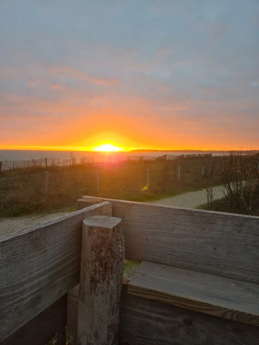 Le soleil vient de se coucher face au Sunset : ciel orangé sur l’horizon sous de grands nuages gris, au premier plan la terrasse en bois, puis la dune et ses ganivelles, le chemin de sable vers la plage et la mer