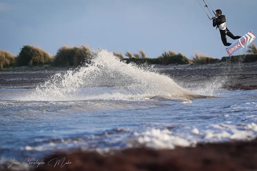 Un kitesurfeur en plein saut au-dessus d’une gerbe d’eau, devant une côte basse couverte d’herbes hautes